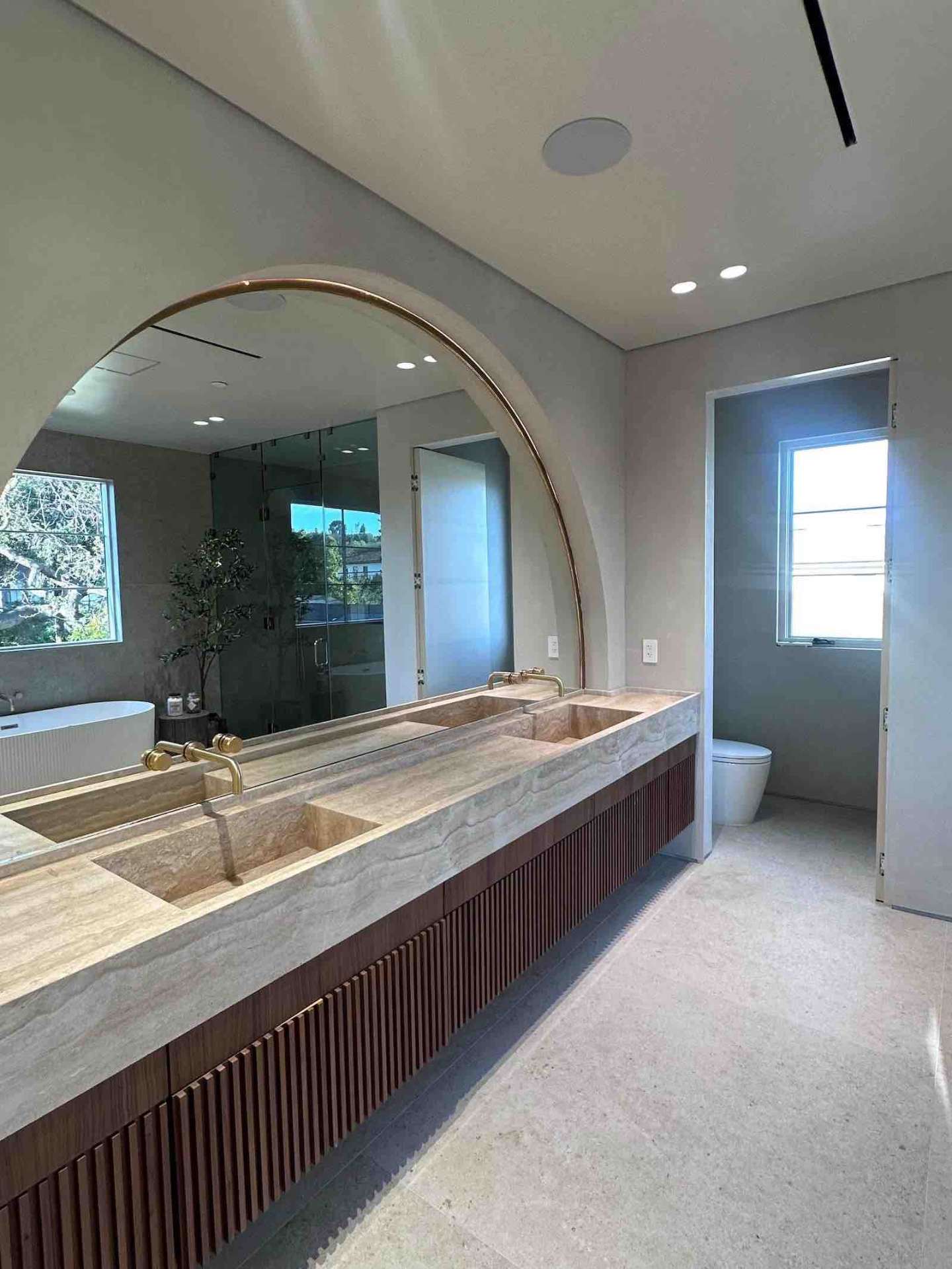 Encino double vanity with travertine integrated sinks, brass faucets, and arch mirror